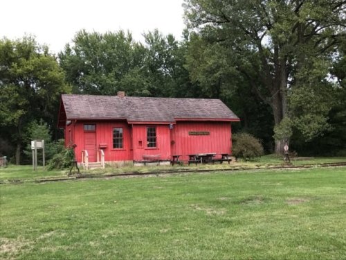 A red wooden building surrounded by greenery, with a grassy area in front and trees in the background.