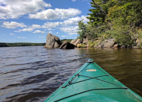 A green kayak on calm water, with rocky shore and trees under a blue sky with fluffy clouds.