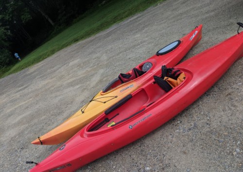 Two kayaks, one red and one orange, resting on a gravel surface with trees in the background.