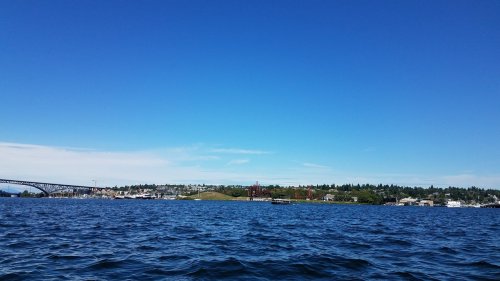 A clear blue sky above a calm water body, with a distant shoreline featuring buildings and greenery.