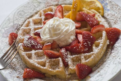 A golden waffle topped with fresh strawberries, whipped cream, and a slice of orange, served on a decorative plate.