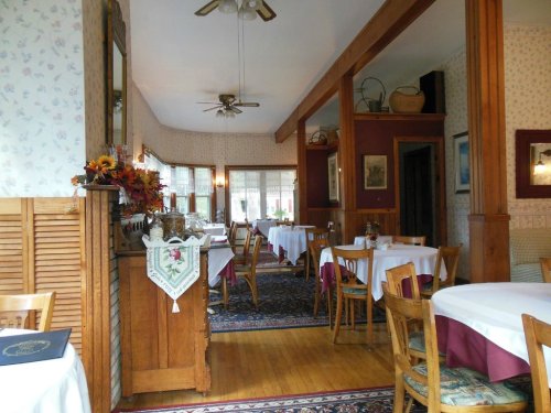 Cozy restaurant interior with wooden furniture, white tablecloths, and floral wallpaper, featuring a warm, inviting atmosphere.