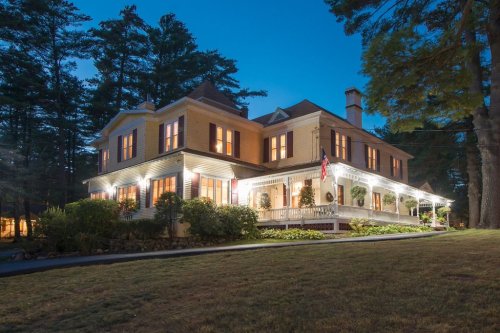 A charming two-story house with a wraparound porch, illuminated at dusk, surrounded by trees and a well-kept lawn.