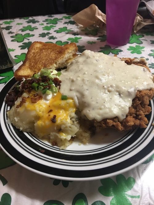A plate of fried chicken smothered in gravy, served with loaded mashed potatoes and a slice of toast.