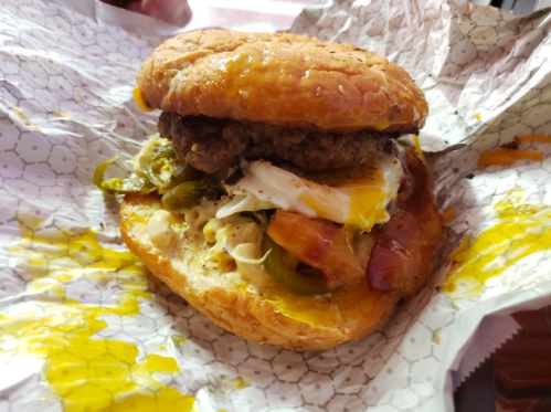 A close-up of a burger with a beef patty, fried egg, jalapeños, and melted cheese on a toasted bun, wrapped in paper.
