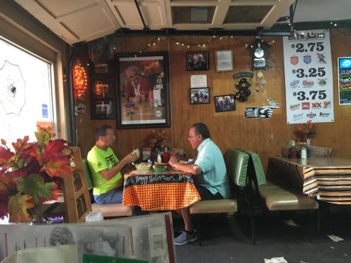 Two men sit at a table in a cozy diner, enjoying meals surrounded by warm decor and a menu on the wall.
