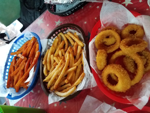 Three baskets of fried snacks: sweet potato fries, regular fries, and onion rings, served on a red table with a star pattern.
