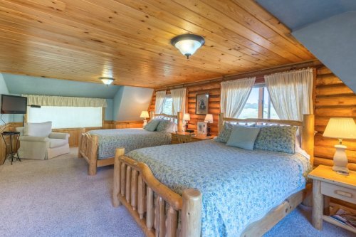 Cozy log cabin bedroom with two beds, wooden ceiling, and large windows, featuring soft lighting and rustic decor.