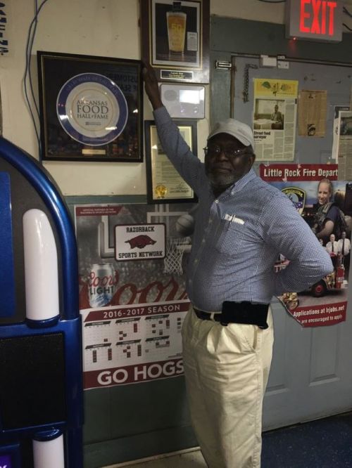 An older man in a blue shirt stands next to framed awards on a wall in a casual setting.