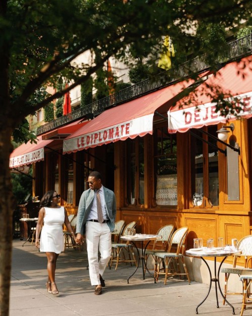A couple walks hand in hand outside a charming restaurant with outdoor seating and colorful awnings.