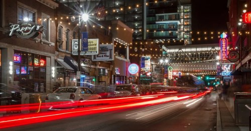 A vibrant city street at night, illuminated by neon signs and string lights, with light trails from passing cars.