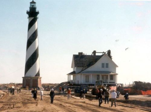 A black-and-white striped lighthouse next to a house under construction, with people walking on a sandy path.