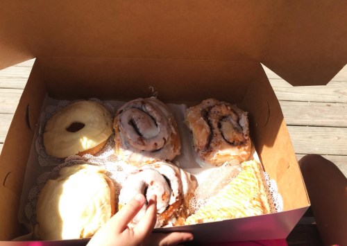 A box of assorted pastries, including glazed donuts and cinnamon rolls, with a hand reaching for one.