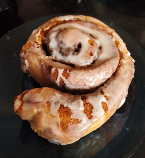 A freshly baked cinnamon roll drizzled with creamy icing, sitting on a dark plate.