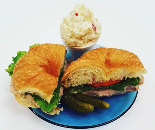 A croissant sandwich filled with greens and tomato, served with pickles and a bowl of creamy potato salad.