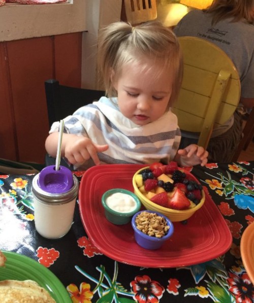 A young child sits at a table, enjoying a colorful fruit bowl with yogurt and granola, surrounded by a vibrant tablecloth.