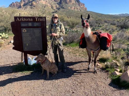 A man with a beard stands by an Arizona Trail sign, accompanied by a dog and a llama with packs. Desert landscape in background.