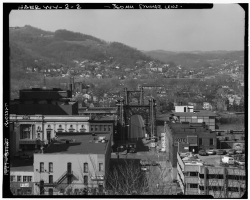 A black and white aerial view of a cityscape featuring a bridge, buildings, and hills in the background.