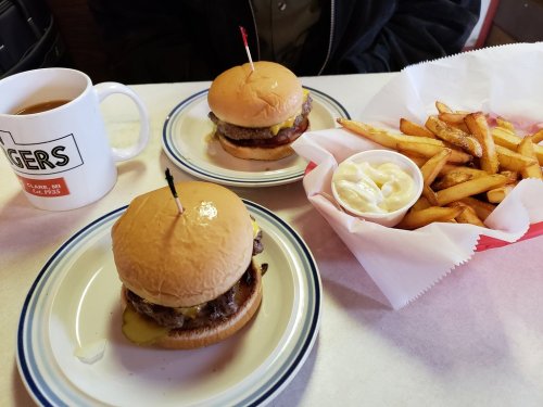 Two cheeseburgers with pickles and fries served with a cup of coffee on a table.
