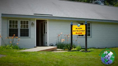 A light gray building with a sign indicating the main entrance, surrounded by green grass and orange flowers.