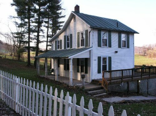 A white two-story house with a green metal roof, surrounded by trees and a white picket fence.