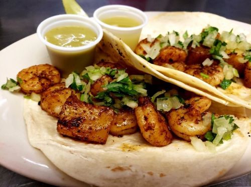 Two shrimp tacos on a plate, topped with onions and cilantro, served with two cups of green sauce.