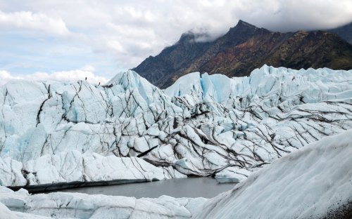 A glacier with blue ice and dark streaks, surrounded by mountains under a cloudy sky. A small lake is visible nearby.