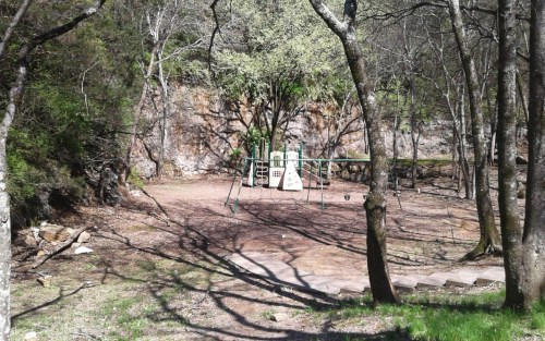 A deserted playground surrounded by trees and rocky terrain, with swings and a slide in a natural setting.