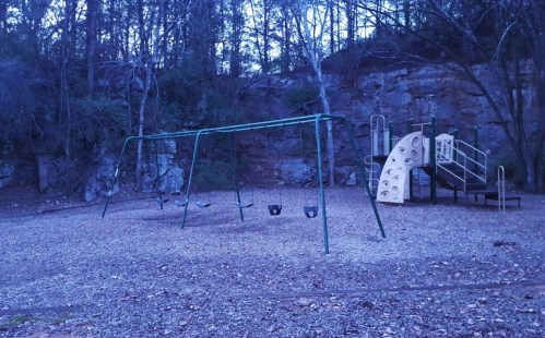 A playground at dusk featuring swings and a climbing structure, surrounded by trees and rocky terrain.