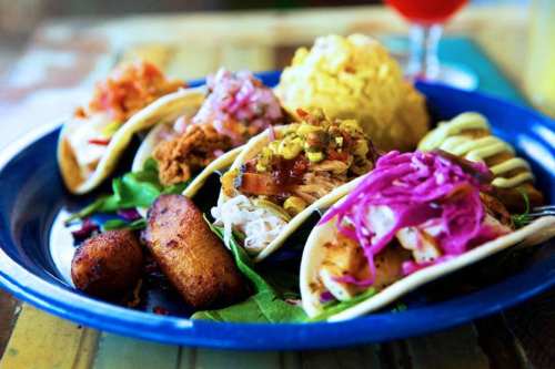 A colorful plate featuring three tacos, rice, and fried plantains, garnished with fresh vegetables.