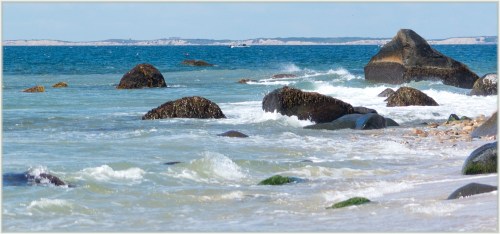 Waves gently crash against rocky shorelines under a clear blue sky, with distant hills visible on the horizon.