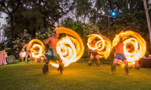 Fire dancers perform in a lush outdoor setting, creating swirling patterns of flames against a twilight backdrop.