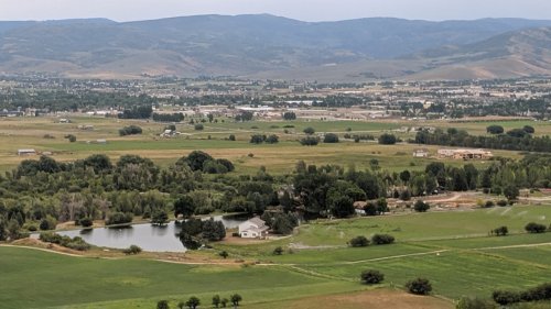 A scenic view of a green landscape with a pond, houses, and mountains in the background under a cloudy sky.