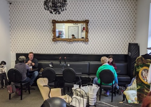 A cozy café interior with black seating, a chandelier, and patrons engaged in conversation at tables.