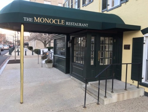 Entrance of The Monocle Restaurant with a green awning and steps leading up to the door, set in a city street.