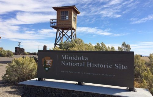Sign for Minidoka National Historic Site with a watchtower in the background against a blue sky and wispy clouds.
