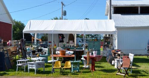 A white tent displays various vintage furniture and decor items on a grassy area beside a barn.