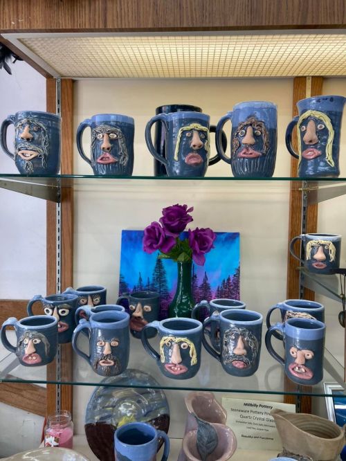 A display of unique ceramic mugs with face designs, arranged on a shelf with purple flowers and a colorful painting in the background.