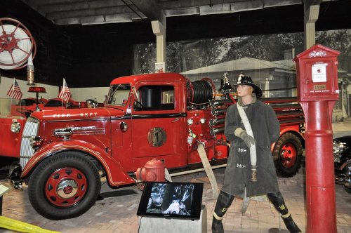 A vintage red fire truck displayed in a museum, with a firefighter mannequin and historical backdrop.