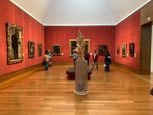 A gallery with red walls, featuring paintings and a central marble sculpture, with visitors exploring the space.