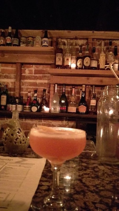 A cocktail sits on a bar counter with a rustic brick wall and shelves filled with various bottles in the background.
