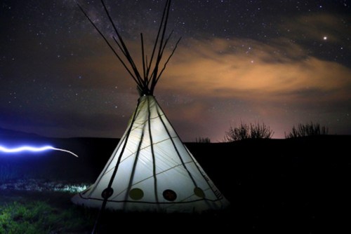 A lit teepee under a starry night sky, with clouds and distant lights in the background.