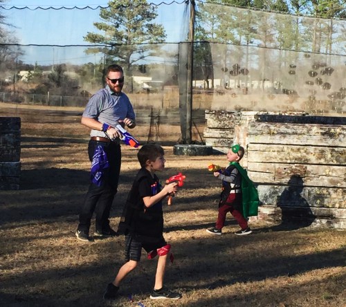 A man and two children play with toy guns in an outdoor setting, surrounded by wooden barriers and trees.