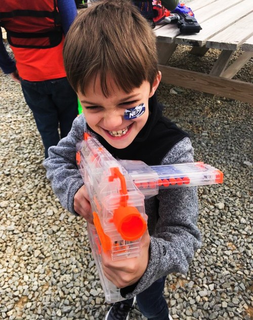 A young boy grins while holding a toy blaster, wearing a sticker on his cheek, outdoors with friends in the background.