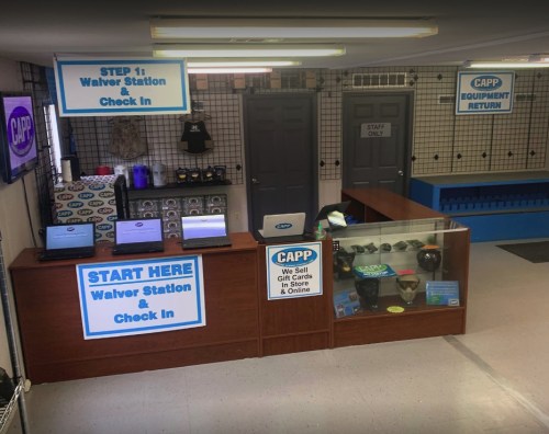 Interior of a check-in area with signs for waivers, equipment return, and gift cards, featuring computers and display cases.