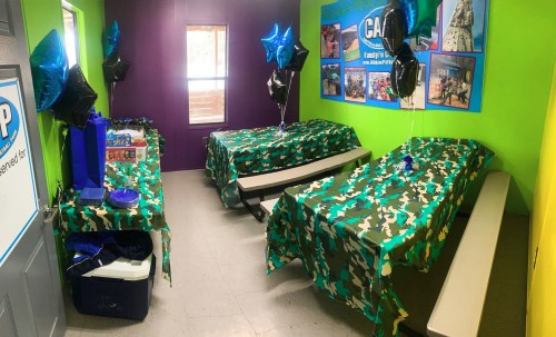 A colorful party room with camo tablecloths, balloons, and decorations, featuring two tables and a cooler.