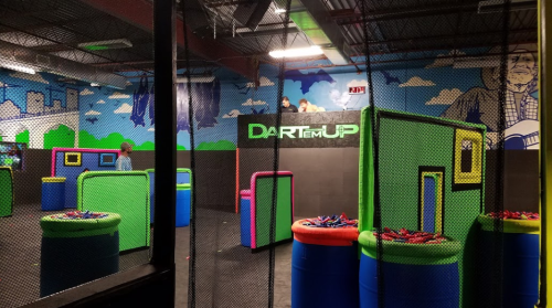 Indoor dart arena with colorful barriers and a netted area, featuring a mural of clouds and a cityscape on the walls.