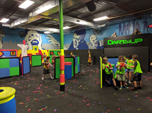 A vibrant indoor play area with kids in neon shirts playing with foam dart blasters among colorful obstacles.