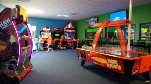 A colorful arcade room with various games, including racing and air hockey, and children playing in the background.