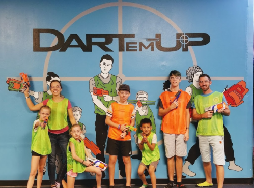 A group of children and adults in colorful vests pose with toy blasters in front of a Dartem Up mural.
